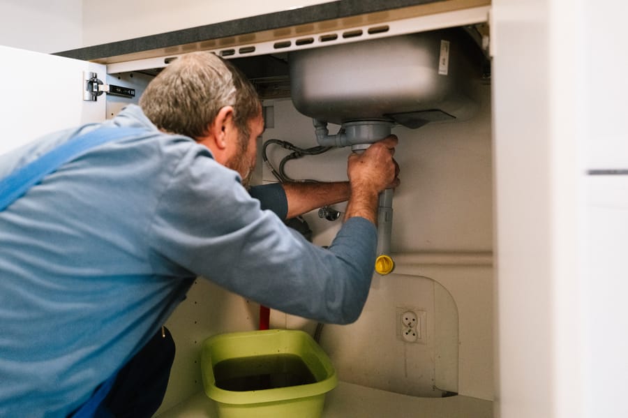 Plumber inspecting drain and sewer piping beneath a kitchen sink.